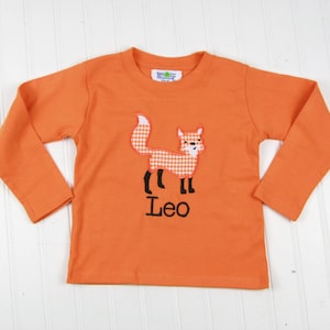 May include: Orange long-sleeved shirt with a fox applique and the name "Leo" embroidered on the front.