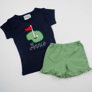 May include: A navy blue short-sleeved t-shirt with a green and white golf course design and the name "Annie" embroidered on the front. The shirt is paired with green and white gingham shorts.