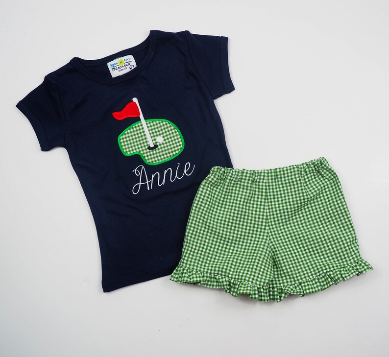 May include: A navy blue short-sleeved t-shirt with a green and white golf course design and the name "Annie" embroidered on the front. The shirt is paired with green and white gingham shorts.