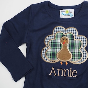 May include: A navy blue long-sleeved shirt with a brown and green embroidered turkey design. The shirt has the text "Soda City Sewing" and "Size 24M" on a yellow tag. The name "Annie" is embroidered below the turkey.