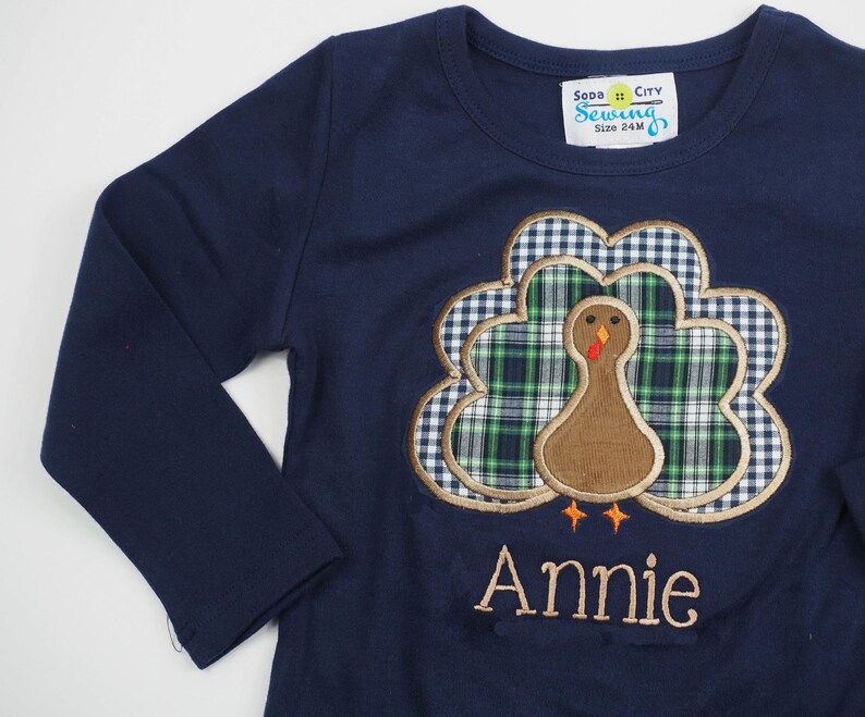 May include: A navy blue long-sleeved shirt with a brown and green embroidered turkey design. The shirt has the text "Soda City Sewing" and "Size 24M" on a yellow tag. The name "Annie" is embroidered below the turkey.