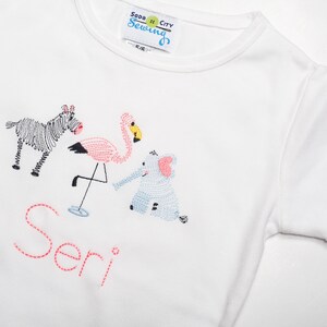 K&ouml;nnte beinhalten: Wei&szlig;es T-Shirt mit gestickten Tieren, darunter ein Zebra, ein Flamingo und ein Elefant. Der Name "Seri" ist in Pink unter den Tieren gestickt.