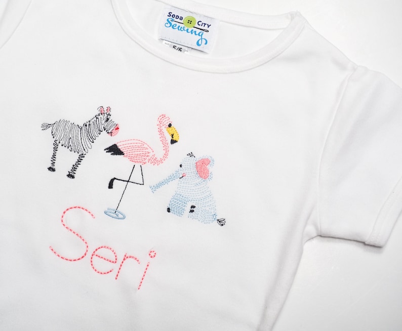 K&ouml;nnte beinhalten: Wei&szlig;es T-Shirt mit gestickten Tieren, darunter ein Zebra, ein Flamingo und ein Elefant. Der Name "Seri" ist in Pink unter den Tieren gestickt.