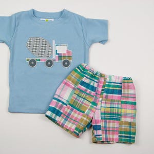 Op de afbeelding: Een lichtblauw T-shirt met korte mouwen met een grijze en witte cementtruck-applicatie. Het shirt wordt gecombineerd met geruite shorts in roze, groen en blauw.