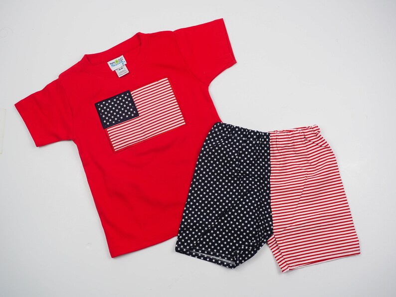May include: Red short-sleeved t-shirt with a blue and red American flag design. Matching red, white, and blue patterned shorts.