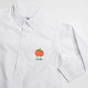 May include: White button-down shirt with a pocket featuring an embroidered orange tomato with a green stem. The name "Jude" is embroidered below the tomato. The shirt has long sleeves and a button-down collar.