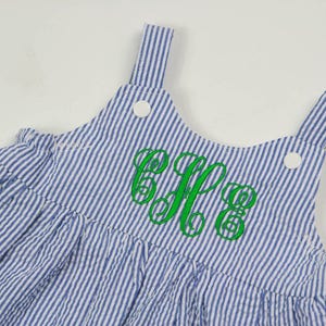 K&ouml;nnte beinhalten: Ein blaues und wei&szlig; gestreiftes Latzkleid mit einem gr&uuml;nen gestickten Monogramm. Das Kleid hat einen R&uuml;schen-Saum und verstellbare Tr&auml;ger.