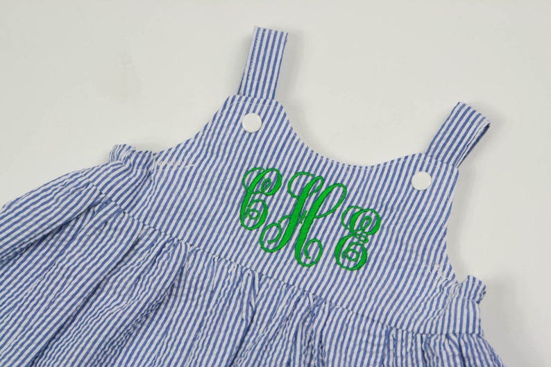 K&ouml;nnte beinhalten: Ein blaues und wei&szlig; gestreiftes Latzkleid mit einem gr&uuml;nen gestickten Monogramm. Das Kleid hat einen R&uuml;schen-Saum und verstellbare Tr&auml;ger.