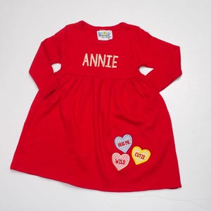 Könnte beinhalten: Rotes Langarmkleid mit einem weißen gestickten Namen "ANNIE" auf der Vorderseite. Das Kleid hat drei gestickte Bonbonherzen mit den Worten "HUG ME", "WILD" und "CUTIE" darauf.