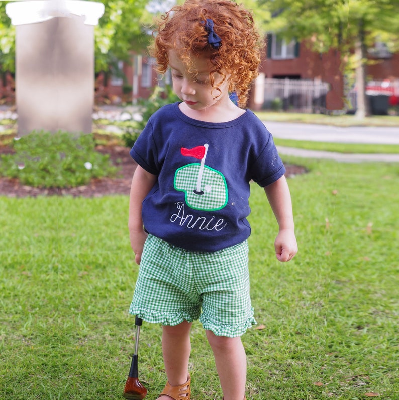 May include: A young girl wearing a navy blue t-shirt with a green and white embroidered golf course design and the name "Annie" on it. She is also wearing green and white gingham shorts with a ruffle trim.