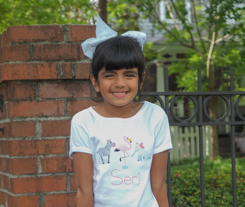 K&ouml;nnte beinhalten: Ein junges M&auml;dchen mit einer blauen Schleife im Haar tr&auml;gt ein wei&szlig;es T-Shirt mit einem Flamingo-, Zebra- und Elefantenmotiv. Das Shirt tr&auml;gt den Schriftzug "Seri" unten.