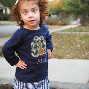 May include: A young girl with curly red hair wears a navy blue long-sleeved shirt with a plaid turkey applique and the name "Annie" embroidered on the front. She is also wearing a blue and white gingham skirt.