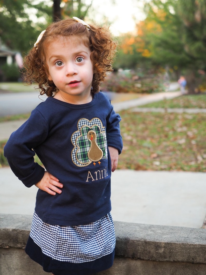 May include: A young girl with curly red hair wears a navy blue long-sleeved shirt with a plaid turkey applique and the name "Annie" embroidered on the front. She is also wearing a blue and white gingham skirt.