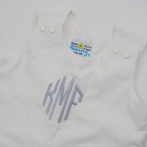 Puede incluir: Pantalón vaquero blanco con un monograma bordado en gris "KMF" y una etiqueta azul y blanca que dice "Soda City Sewing Talla 9M".