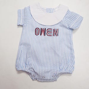 Pode incluir: Um macacão de bebê com gola branca e mangas curtas. O macacão é listrado em azul claro e branco com o nome "OWEN" bordado em vermelho e azul marinho. O macacão tem fechos de pressão na parte inferior.