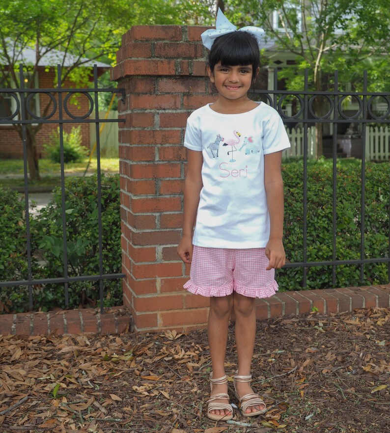 K&ouml;nnte beinhalten: Ein junges M&auml;dchen tr&auml;gt ein wei&szlig;es T-Shirt mit einem gestickten Design von Tieren und dem Namen "Seri" darauf. Sie tr&auml;gt auch rosa und wei&szlig;e Gingham-Shorts mit R&uuml;schenbesatz und eine blaue Schleife im Haar.