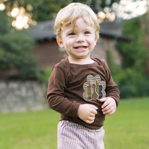 May include: A young child wearing a brown long-sleeved shirt with a colorful turkey applique. The shirt has a brown collar and a brown trim around the applique. The child is wearing brown and white plaid pants.