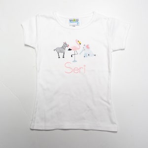 K&ouml;nnte beinhalten: Wei&szlig;es T-Shirt mit einem Flamingo-, Zebra- und Elefantenmotiv. Der Name "Seri" ist in Pink unter den Tieren gestickt.