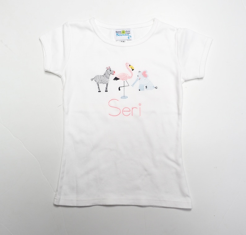 K&ouml;nnte beinhalten: Wei&szlig;es T-Shirt mit einem Flamingo-, Zebra- und Elefantenmotiv. Der Name "Seri" ist in Pink unter den Tieren gestickt.