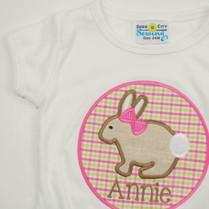Op de afbeelding: Wit T-shirt met een roze en groene ruitjescirkel met een bruin konijn met een roze strik en de naam "Annie" geborduurd aan de onderkant.