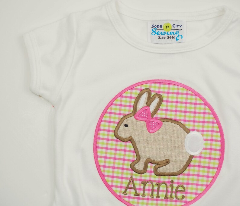 Op de afbeelding: Wit T-shirt met een roze en groene ruitjescirkel met een bruin konijn met een roze strik en de naam "Annie" geborduurd aan de onderkant.