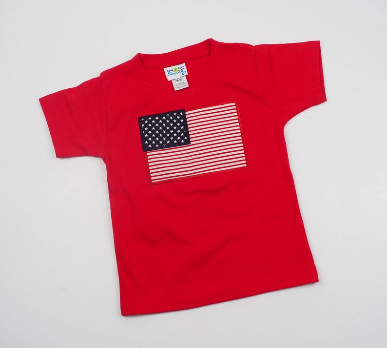 May include: Red short-sleeved t-shirt with an embroidered American flag on the front. The flag is white and blue with red stripes.