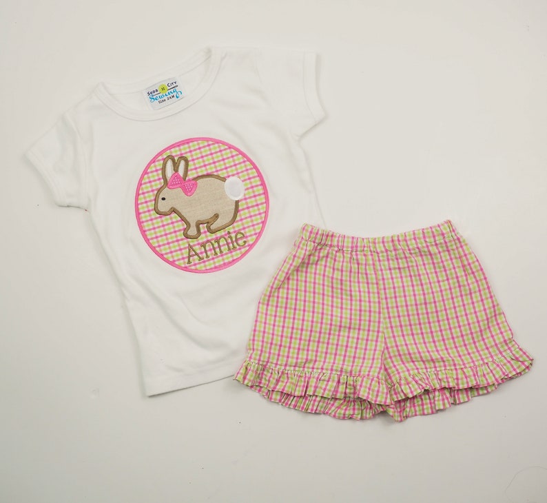 Op de afbeelding: Een wit T-shirt met korte mouwen met een roze en groene ruitvormige cirkelapplicatie met een bruin konijn met een roze strik. Het shirt is gepersonaliseerd met de naam "Annie". De outfit wordt gecombineerd met roze en groene ruitvormige shorts met een gerimpelde zoom.
