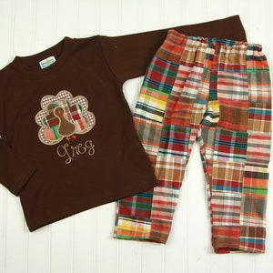 Könnte beinhalten: Ein braunes Langarmshirt mit einem gestickten Truthahn-Motiv und dem Namen "Greg" darunter. Das Shirt wird mit karierten Hosen mit Patchwork-Muster in Rot-, Braun- und Weißtönen kombiniert.