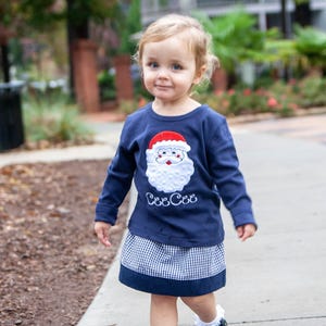 Könnte beinhalten: Ein kleines Kind trägt ein marineblaues Langarmshirt mit einem weißen Weihnachtsmann-Gesicht und dem Text "Cee Cee" auf der Vorderseite. Das Kind trägt außerdem einen marineblauen und weißen Gingham-Rock.
