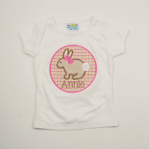 Op de afbeelding: Wit T-shirt met een roze en groene gingham cirkel met een bruin konijn met een roze strik en de naam "Annie" erop geborduurd.