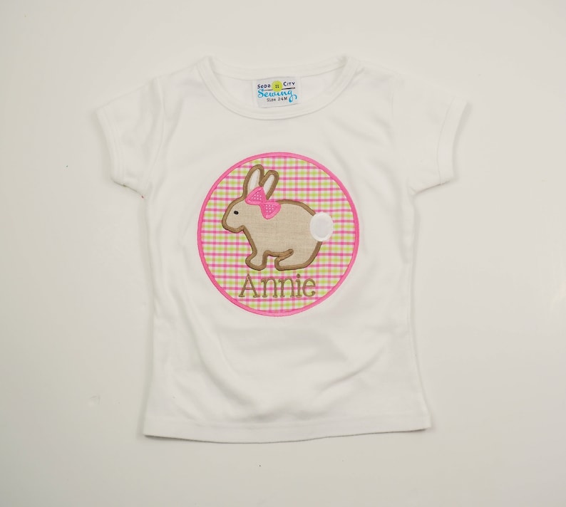 Op de afbeelding: Wit T-shirt met een roze en groene gingham cirkel met een bruin konijn met een roze strik en de naam "Annie" erop geborduurd.