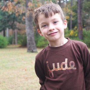 May include: A young person wearing a brown long-sleeved shirt. The shirt features the name "jude" embroidered in colorful thread. The person is smiling, with a blurred outdoor background of trees and foliage.