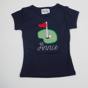 May include: A navy blue t-shirt with a green and white checkered golf green applique and a red flag. The name "Annie" is embroidered in white below the applique. The shirt has a white tag with the text "Seashore City Sewing Size 2T".