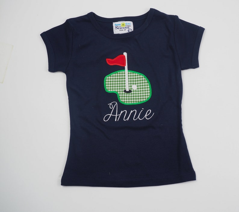 May include: A navy blue t-shirt with a green and white checkered golf green applique and a red flag. The name "Annie" is embroidered in white below the applique. The shirt has a white tag with the text "Seashore City Sewing Size 2T".