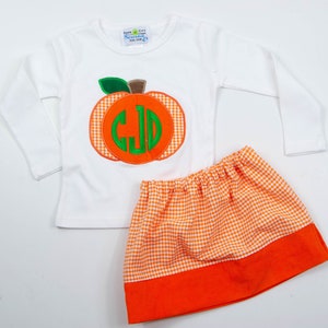 Op de afbeelding: Een wit longsleeve shirt met een oranje en groene pompoen-applicatie met een monogram. Het shirt wordt gecombineerd met een oranje en witte gingham rok met een oranje band aan de onderkant.