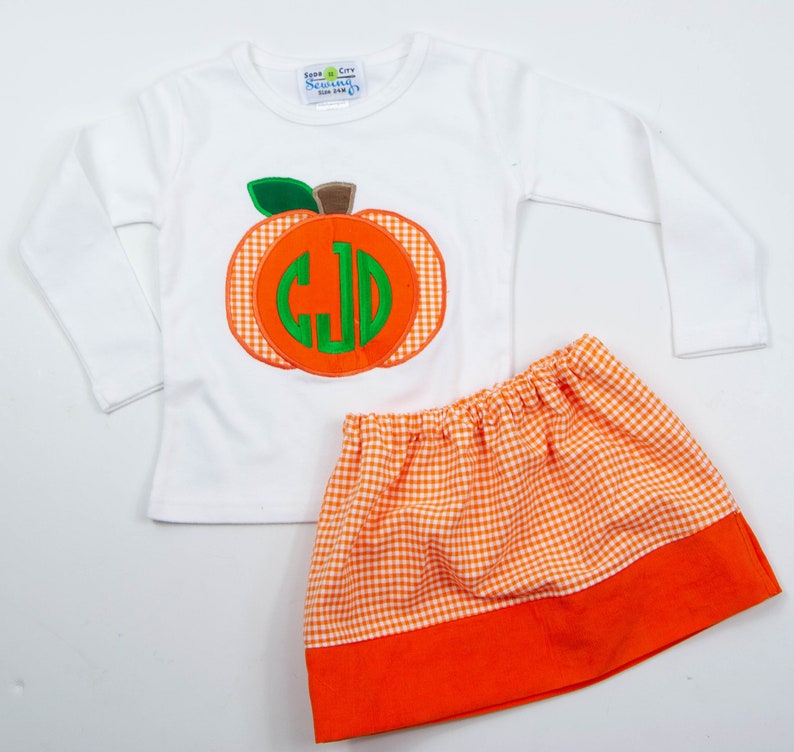 Op de afbeelding: Een wit longsleeve shirt met een oranje en groene pompoen-applicatie met een monogram. Het shirt wordt gecombineerd met een oranje en witte gingham rok met een oranje band aan de onderkant.