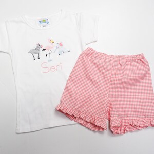 K&ouml;nnte beinhalten: Wei&szlig;es T-Shirt mit kurzen &Auml;rmeln mit einem pinkfarbenen Flamingo-, Zebra- und Elefanten-Motiv und dem Namen "Seri", der auf der Vorderseite gestickt ist. Das T-Shirt wird mit pinkfarbenen und wei&szlig;en Gingham-Shorts mit R&uuml;schen am Saum kombiniert.