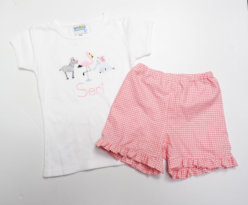 K&ouml;nnte beinhalten: Wei&szlig;es T-Shirt mit kurzen &Auml;rmeln mit einem pinkfarbenen Flamingo-, Zebra- und Elefanten-Motiv und dem Namen "Seri", der auf der Vorderseite gestickt ist. Das T-Shirt wird mit pinkfarbenen und wei&szlig;en Gingham-Shorts mit R&uuml;schen am Saum kombiniert.
