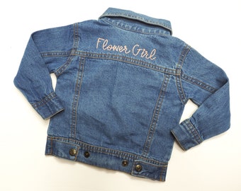Könnte beinhalten: Eine blaue Jeansjacke mit einer rosa gestickten Aufschrift auf der Rückseite, die "Flower Girl" lautet.