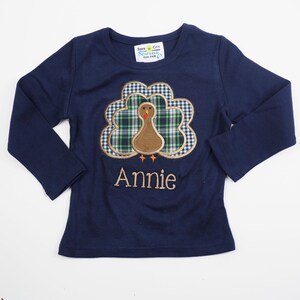 May include: Navy blue long-sleeved shirt featuring a personalised embroidered turkey design. The turkey is made of plaid fabric with a brown head and a white and blue checked background. The name "Annie" is embroidered below the turkey.