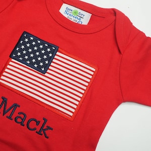 May include: Red baby onesie with a blue embroidered American flag and the name "Mack" in blue embroidery.