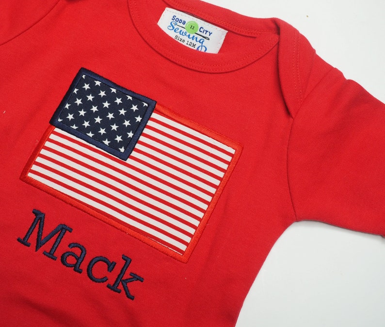 May include: Red baby onesie with a blue embroidered American flag and the name "Mack" in blue embroidery.