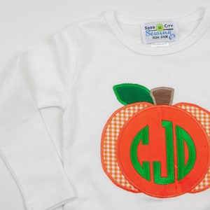 Op de afbeelding: Witte longsleeve met een groene en oranje pompoen-applicatie. De pompoen heeft een wit en oranje ruitjespatroon en een groen monogram in het midden. Het shirt heeft een label met de tekst "Soda City Sewing Maat 24M".