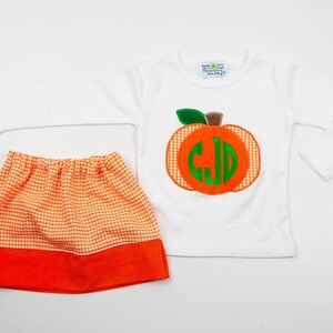 Op de afbeelding: Een wit longsleeve shirt met een groene en oranje pompoen applique met een monogram. Het shirt wordt gecombineerd met een wit en oranje gingham rok met een oranje onderkant.