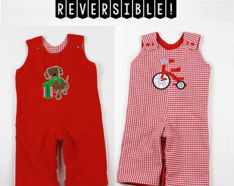 Puede incluir: Pantalones de peto reversibles para bebé en rojo y blanco. Un lado presenta una tela roja con un perro marrón bordado que sostiene un regalo verde. El otro lado presenta un patrón de cuadros rojos y blancos. Los pantalones de peto tienen una pancarta "REVERSIBLE!" en la parte superior.