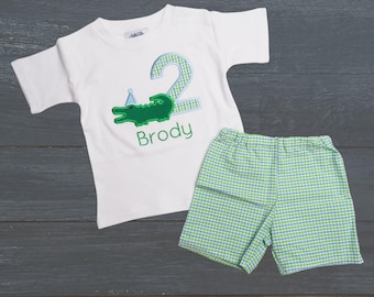 May include: A white short-sleeved t-shirt with a green alligator wearing a party hat and a blue and white checkered number two. The shirt also has the name "Brody" embroidered on it. The shirt is paired with green and blue checkered shorts.
