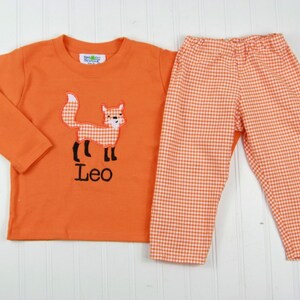 May include: Orange long-sleeve shirt with a fox applique and the name "Leo" embroidered on the front. Matching orange and white gingham pants.
