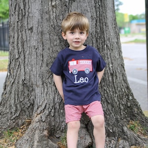 May include: A child's outfit featuring a navy blue t-shirt with a red and white gingham fire engine and the name "Leo". The outfit includes red and white gingham shorts. The child is standing by a tree.