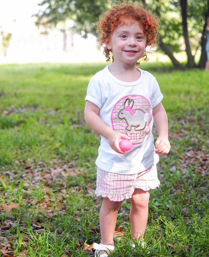 Op de afbeelding: Een jong meisje met krullend rood haar draagt een wit T-shirt met een roze konijnenapplicatie en de naam "Annie" geborduurd erop. Ze draagt ook roze en witte geruite shorts en houdt een roze paasei vast.
