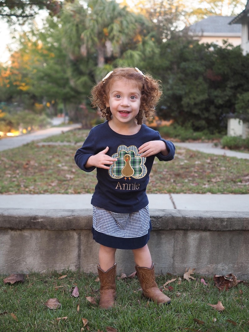 May include: A young girl wearing a blue long-sleeved shirt with a turkey applique and the name "Annie" embroidered on it. She is also wearing a plaid skirt and brown cowboy boots.